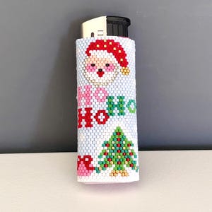 Op de afbeelding: Een aansteker met een kralenhoes op een lichtblauwe achtergrond, voorzien van een gepixeld kerstmangezicht met een rode muts, een groene kerstboom en de rood-groene tekst "HO HO HO" die verticaal op de voorzijde is geplaatst.