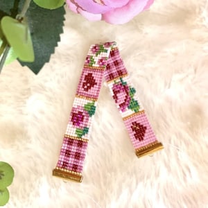 Op de afbeelding: Een roze en witte kralen armband met een bloemenpatroon. De armband is gemaakt van kleine kralen en heeft een herhalend patroon van roze bloemen en groene bladeren.
