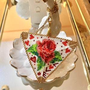 May include: A triangular pendant necklace with a beaded design featuring a red rose and hearts. The pendant is made of gold-colored metal and hangs from a gold chain.