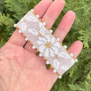 Op de afbeelding: Een kralenarmband met een wit en lichtbruin bloemmotief. De armband is afgewerkt met goudkleurige kralen en kleine witte parels. De armband wordt in een hand gehouden tegen een achtergrond van groen gebladerte.