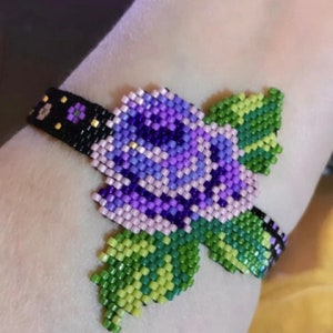 Peut inclure: Un bracelet avec une fleur en perles violettes et roses et des feuilles vertes. Le bracelet est porté au poignet.