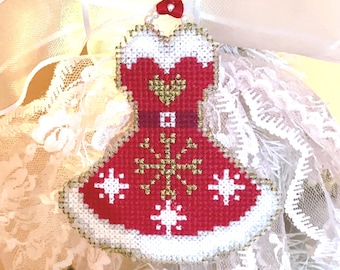 May include: A red and white Christmas tree ornament shaped like a dress, with gold accents and snowflake details. The ornament is hanging from a red ribbon. The background includes white lace and a glittery gold star.