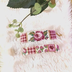 Könnte beinhalten: Zwei Perlenarmbänder mit rosa und weißen Karomustern und Blumendesigns. Die Armbänder liegen auf einer weißen, flauschigen Oberfläche.