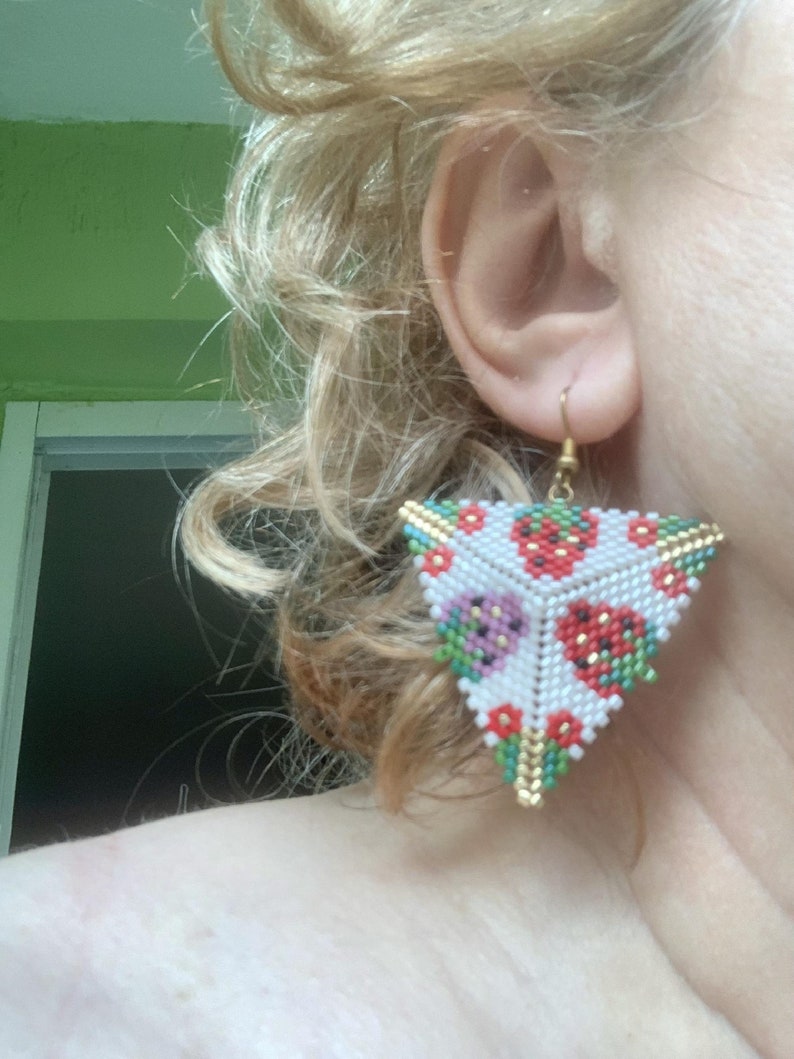 May include: Triangle-shaped earrings with a white background and a pattern of red, green, and purple strawberries. The earrings are made of beads and have a gold-colored wire.