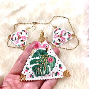 May include: A beaded jewelry set featuring a triangular pendant and matching earrings. The white background is adorned with pink flamingos, green leaves, and gold accents. The set is connected by delicate gold chains.