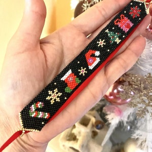 May include: A black beaded bracelet with a red ribbon and a Christmas-themed design. The design includes a stocking, a snowflake, a candy cane, a Christmas tree, a gift box, and a Santa hat.