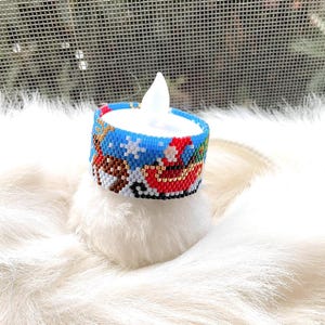 May include: A battery-operated LED candle with a beaded Christmas design. The blue, red, white, and brown beaded band features Santa Claus in a sleigh, snowflakes, and a winter scene. The candle sits on a fluffy white surface.