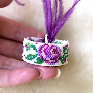 May include: A white beaded ring with a purple and pink floral design. The ring is topped with a purple feather.