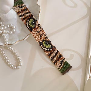 May include: A beaded bracelet with a green, brown, and tan pattern. The bracelet features an eye design. The bracelet is on a white surface with a pearl necklace and a decorative dish.