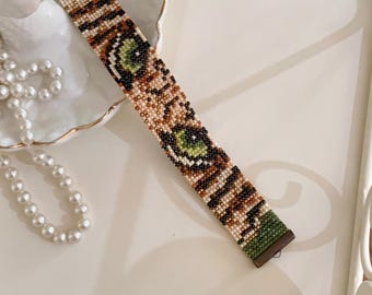 May include: A beaded bracelet with a green, brown, and tan pattern. The bracelet features a design resembling an eye. The bracelet is on a white surface with a pearl necklace and a decorative dish.