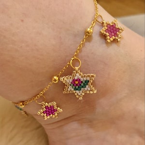 Könnte beinhalten: Goldkette Fußkettchen mit drei sternförmigen Anhängern. Die Anhänger sind aus bunten Perlen gefertigt und haben verschiedene Muster.