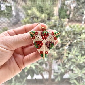 May include: A triangular beaded earring with a floral pattern in red, white, green, and gold. The earring is made of small beads and has a gold hook.