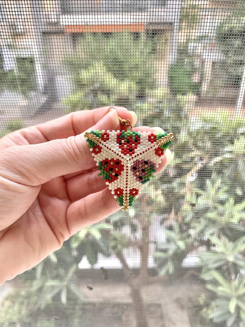 May include: A triangular beaded earring with a floral pattern in red, white, green, and gold. The earring is made of small beads and has a gold hook.