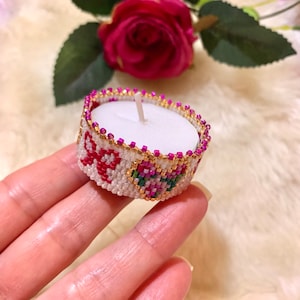 May include: A small white candle holder with a beaded design featuring pink, red, and green flowers. The holder is made of white beads with gold accents.