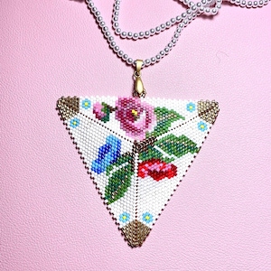 Pode incluir: Um colar de contas com um pingente triangular. O pingente apresenta um design floral com uma rosa rosa, um pássaro azul e bagas vermelhas em um fundo branco. As bordas são adornadas com contas douradas e o colar tem uma corrente de contas brancas.