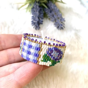 May include: A white beaded bracelet with a floral design featuring purple and green beads. The bracelet is made with small, square beads.
