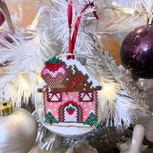 May include: A pink and brown gingerbread house Christmas ornament with a red ribbon and a white frosting roof. The house has a green door and two small green trees on either side.