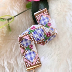 May include: A beaded bracelet with a floral pattern in shades of pink, purple, white, and green. The bracelet has a gold clasp.