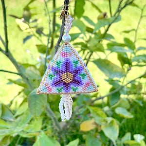 May include: A handcrafted, beaded triangle ornament with a floral design in shades of purple, pink, green, and gold. The ornament is suspended from a dark cord and adorned with a beaded tassel. The background is a blurred view of green foliage.