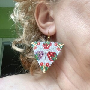 May include: Triangle-shaped beaded earrings with a white background and a pattern of red, green, and purple strawberries. The earrings are made with small beads and have a gold-colored wire.