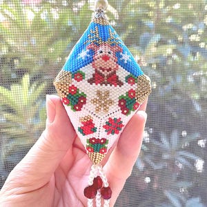 May include: A handmade Christmas ornament in a diamond shape, featuring a reindeer design. The ornament is adorned with red, green, and gold beads, with a beaded tassel of pearls and red beads. The top has a small red bead.