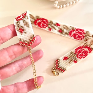 May include: A white beaded choker necklace with red roses and gold accents. The necklace has a gold chain clasp.