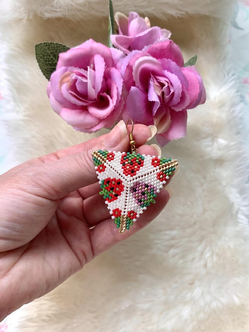 May include: Triangle-shaped beaded earrings with a white background and a colorful pattern of red, green, and gold beads. The pattern resembles strawberries.