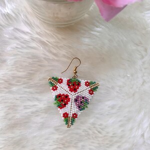 May include: A pair of triangle-shaped earrings with a white background and a beaded design of red strawberries, green leaves, and purple flowers.