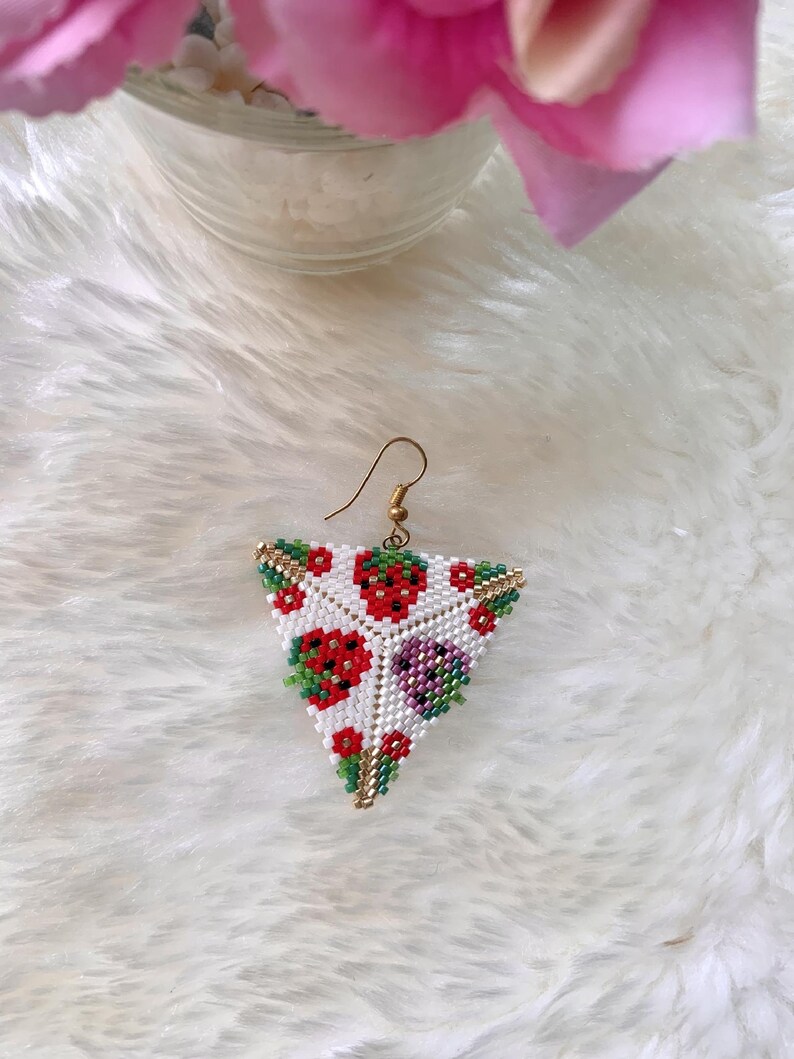 May include: A pair of triangle-shaped earrings with a white background and a beaded design of red strawberries, green leaves, and purple flowers.