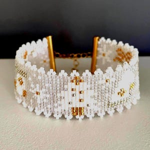Puede incluir: Una pulsera ancha de cuentas con un patrón geométrico en blanco y oro metálico. La pulsera está hecha de pequeñas cuentas uniformes con un borde festoneado en la parte superior e inferior, e incluye terminales de metal dorado y una cadena.