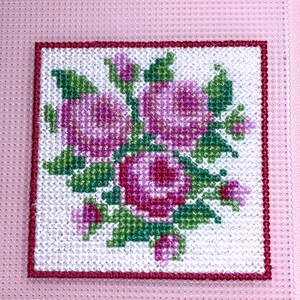 May include: Cross-stitch floral design featuring pink roses and green leaves on a white background, framed by a red border. The needlework is set against a pink, perforated plastic canvas.