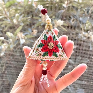 Può includere: Ornamento natalizio fatto a mano a forma triangolare, con un motivo di stella di Natale rossa e foglie verdi su sfondo bianco. L'ornamento è rifinito con oro e decorato con perle e perline, creando una decorazione festiva.