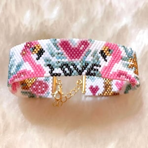 May include: A white beaded bracelet with a pink flamingo design and gold accents. The bracelet has a gold chain clasp.