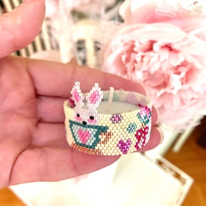 May include: A beaded candle holder with a bunny design. The candle holder is cylindrical, with a cream-colored background and a pattern of pink and purple flowers. A pink bunny with a heart is featured on the front.