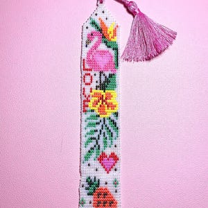 May include: A pink and white beaded bookmark with a flamingo, flowers, a heart, and a pineapple. The bookmark has a pink tassel.