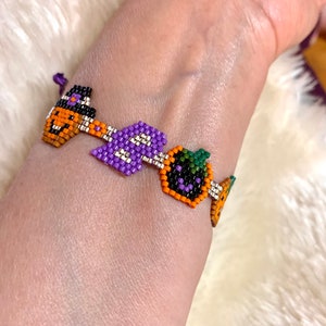 Könnte beinhalten: Ein lila, orange und grünes Perlenarmband mit Halloween-Motiv. Das Armband zeigt einen Hexenhut, einen Kürbis und ein Gespenst.