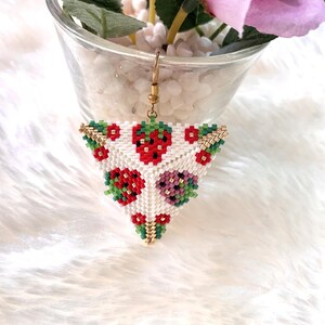 May include: Triangle-shaped beaded earring with a white background and a pattern of red strawberries with green leaves. The earring is made with small beads and has a gold-colored hook.