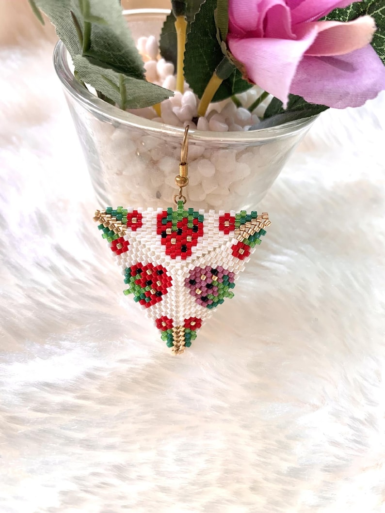 May include: Triangle-shaped beaded earring with a white background and a pattern of red strawberries with green leaves. The earring is made with small beads and has a gold-colored hook.