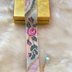 May include: A beaded bracelet with a floral pattern in pink, green, and white. The bracelet has a gold chain and a light blue pearl charm.