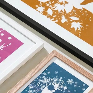 K&ouml;nnte beinhalten: Gerahmte Kunstdrucke mit botanischen und winterlichen Motiven. Die Drucke zeigen wei&szlig;e Silhouetten vor lebendigen Hintergr&uuml;nden in Pink, Blau und Orange. Die Rahmen sind wei&szlig;, schwarz und aus Naturholz.