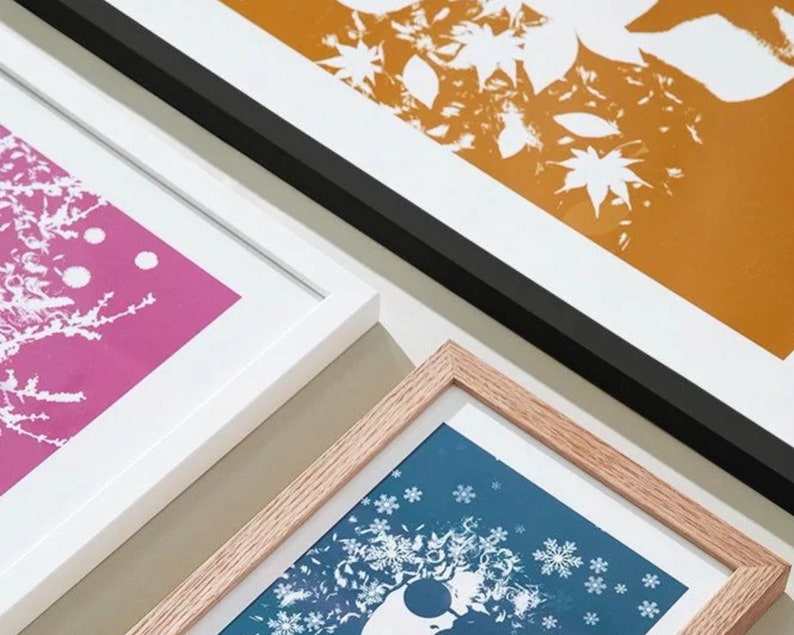K&ouml;nnte beinhalten: Gerahmte Kunstdrucke mit botanischen und winterlichen Motiven. Die Drucke zeigen wei&szlig;e Silhouetten vor lebendigen Hintergr&uuml;nden in Pink, Blau und Orange. Die Rahmen sind wei&szlig;, schwarz und aus Naturholz.