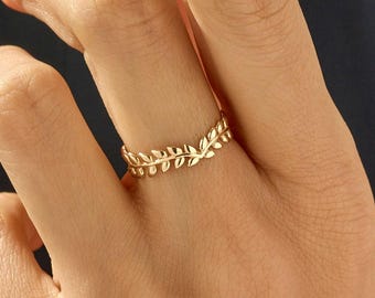 May include: A gold ring featuring a delicate, V-shaped band adorned with a continuous pattern of small, sculpted leaves. The metal has a polished, reflective finish, and the design follows a chevron silhouette that sits centered on the finger.