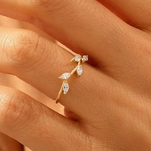 May include: A gold ring featuring a delicate, branch-like band adorned with three small, marquise-cut clear stones arranged in a vine pattern. The metal band has a polished finish and follows a slender, curved design that wraps around the finger.