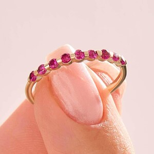 May include: A gold band ring featuring a row of ten small, round, vibrant pink gemstones set along the top half of the band. The ring has a polished metallic finish and a slender profile, showcasing a consistent arrangement of stones across the arc.