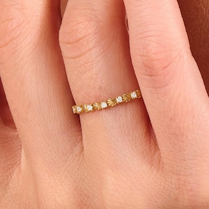 May include: A thin gold band ring featuring a repeating pattern of small, textured shell-like motifs alternating with tiny, clear, round-cut stones set along the entire circumference of the metal band, creating a delicate and uniform appearance.