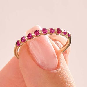 Pode incluir: Um anel de banda dourada com pequenas e vibrantes pedras preciosas rosa. O anel é exibido sobre um fundo rosa suave, mostrando o design delicado do anel e o brilho das pedras. O anel é uma joia.