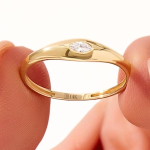 Könnte beinhalten: Ein goldener Ring mit einem Diamanten im Marquiseschliff. Der Ring hat ein glattes, poliertes Band und eine Lünettenfassung für den Diamanten. Der Ring wird zwischen zwei Fingern vor einem weißen Hintergrund gehalten. Der Ring ist mit "14K" gestempelt.