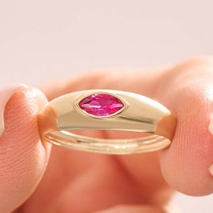 May include: A gold ring with a marquise-cut, bright pink gemstone. The ring is held between two fingers against a soft pink background. The ring's band is smooth and rounded, and the gemstone is set in a bezel.