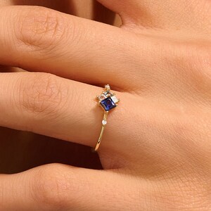 May include: A gold ring featuring a square, dark blue gemstone at the center, accented by smaller white gemstones. The ring has a delicate band and is displayed on a hand with painted nails.