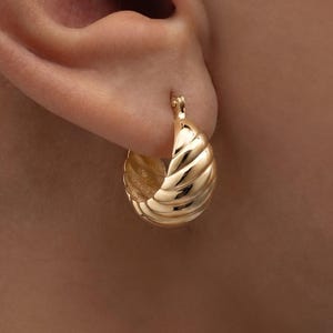 May include: A close-up of a gold hoop earring with a textured, ridged design. The earring is attached to an ear. The earring has a shiny, metallic finish and a classic, timeless style.
